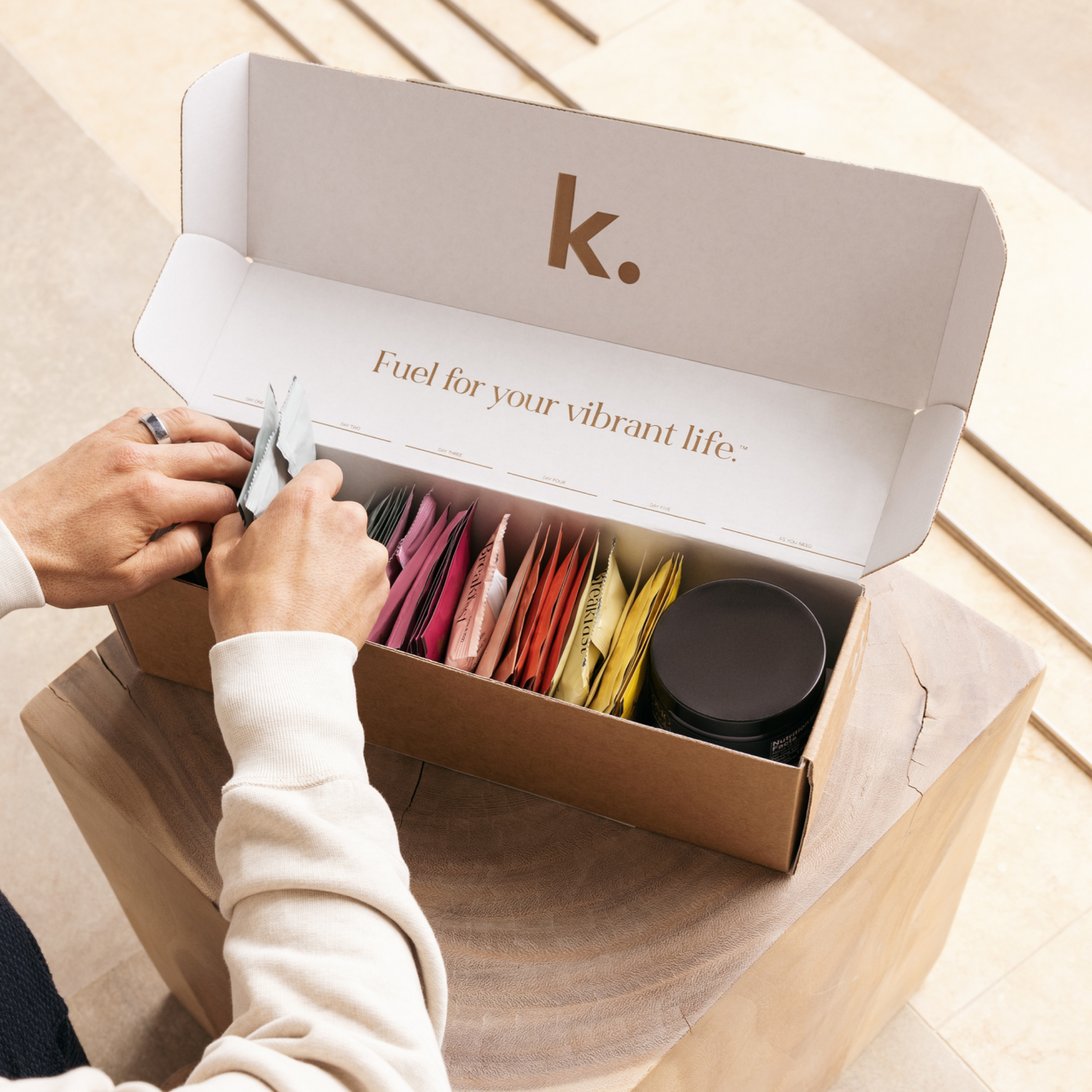 Person opening a box of 'k.' products with colorful packets and a black container.