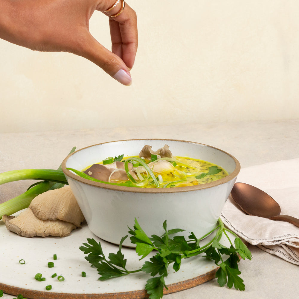 Bowl of healthy soup with herbs and a hand reaching towards it on a light background