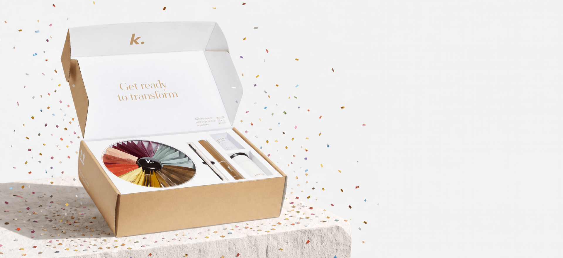An open Kroma Reset box on a light stone surface with confetti scattered around it, containing a circular multicolor makeup wheel, a small brush, and a white booklet. The inside lid shows the brand mark 'k.