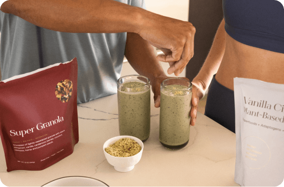 Two people standing at a counter with two glasses of green smoothie, a small bowl of granola, a red bag labeled Super Granola, and a white bag labeled Vanilla Citri Plant-Based powder in a kitchen-like setting.