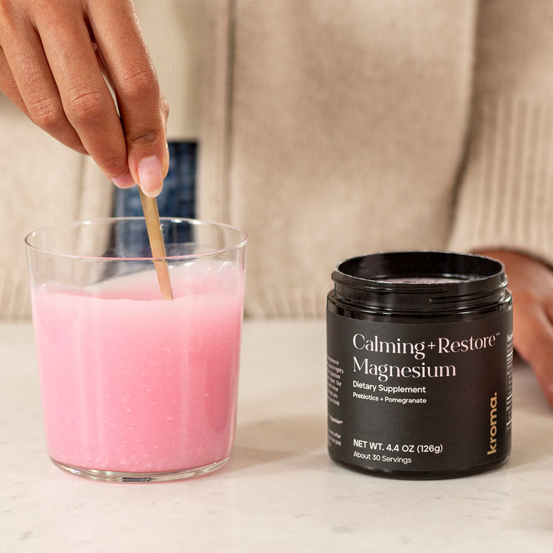 Glass jar with a hand holding a metallic spoon and stirring Kroma’s ,Calming + Restore Magnesium' on a light surface.