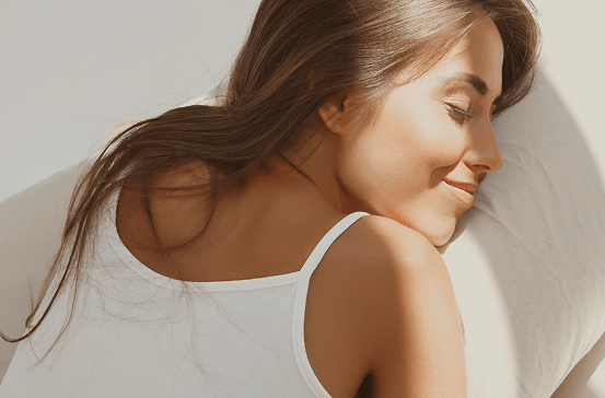 A woman with long brown hair rests her head on a white pillow, wearing a white tank top against a soft white background.